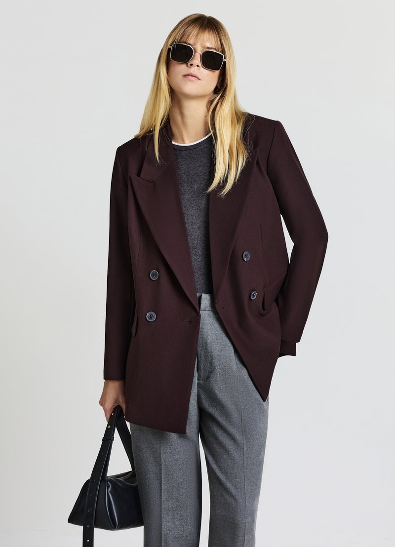 Burgundy double-breasted blazer with notch lapels, black buttons; paired with a gray top and tailored gray trousers; carrying a black handbag.