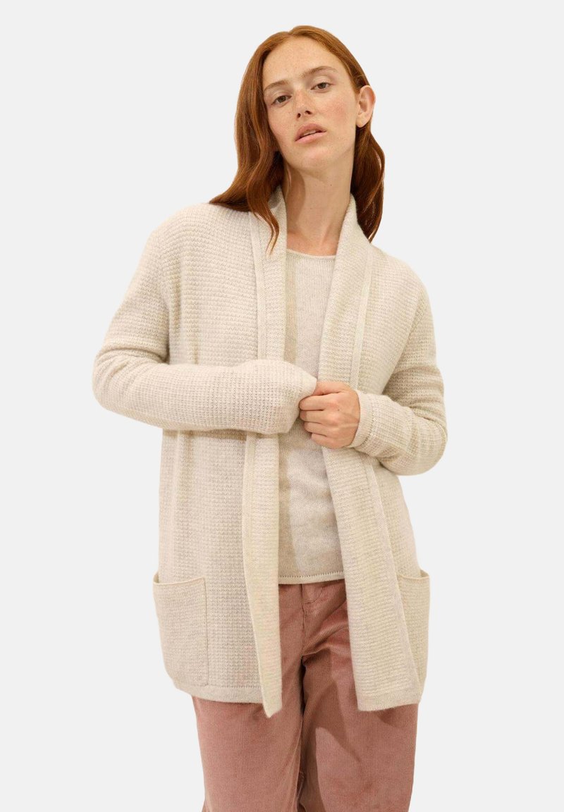 Cardigan de textura beige claro con frente abierto, mangas largas y dos bolsillos frontales. Lleva puesto sobre una blusa beige a juego y pantalones rosa claro.