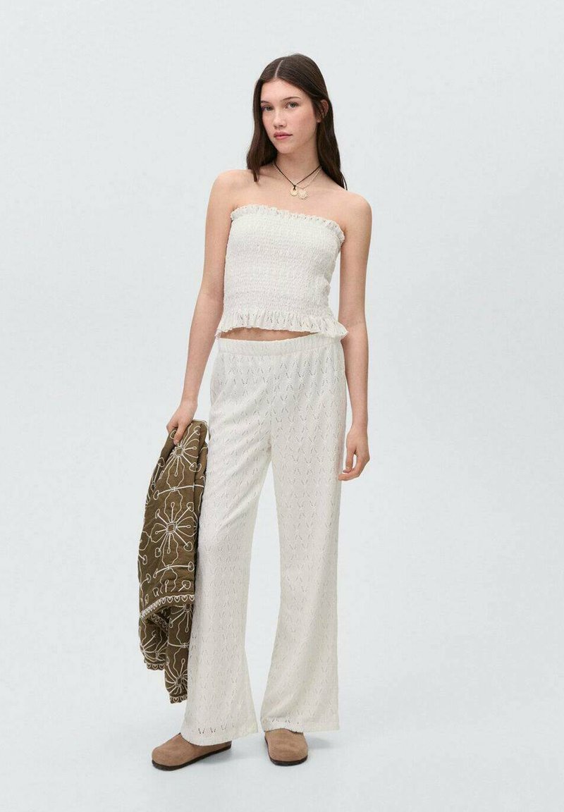 Witte strapless top met rimpelranden, gecombineerd met witte gebloemde wijde broek. Houdt een bruin geborduurd sjaal vast, draagt beige platte schoenen.