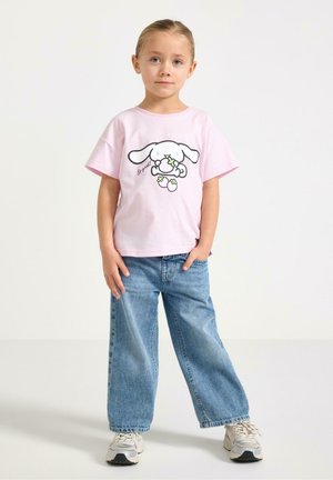 Ung pige stående iført en lyserød T-shirt med tegneseriehund og tekst, løse blå jeans og hvide sneakers på en ensfarvet baggrund.