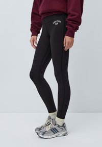 Svarte leggins med høy liv, med logo foran, glatt tekstur og subtile sømdetaljer, sammen med grå joggesko.