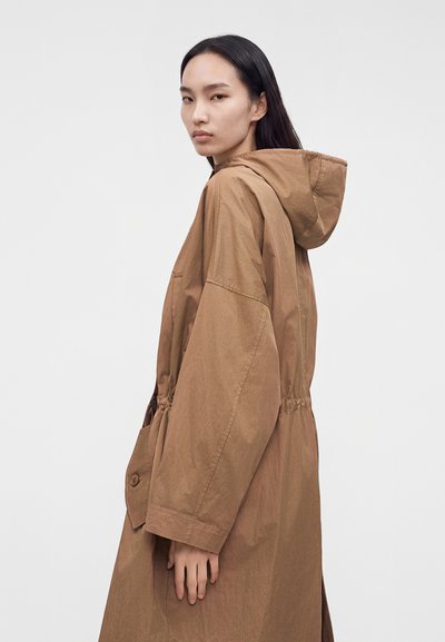 Femme aux longs cheveux foncés portant un manteau marron ample à capuche, de profil sur un fond blanc uni.