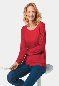 Rotes Langarmoberteil aus weichem Material, mit rundem Halsausschnitt und seitlichen Schlitzen, kombiniert mit blauen Jeans.