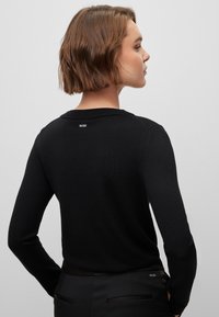 Svart strikkegenser med lange ermer, med ribbestrikket krage og en liten hvit logo på baksiden av halsen. Glatt tekstur og tettsittende design.