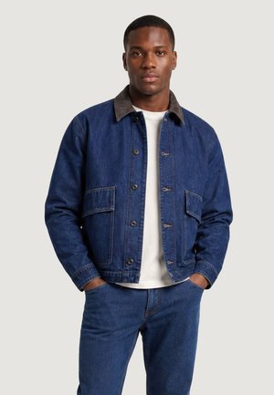 Uomo con giacca di jeans blu con colletto marrone, maglietta bianca e jeans blu, in piedi con le mani nelle tasche su sfondo semplice.