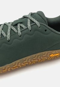 Scarpa sportiva verde scuro con materiale testurizzato, design ondulato sulla parte superiore, lacci grigi e suola in gomma Vibram con motivi geometrici.