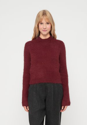 Pull en velours de couleur bordeaux, avec un col haut et des manches longues. Design court, avec une texture douce. Associé à un pantalon sombre à motifs.