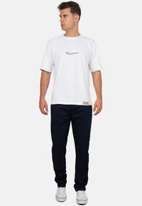 T-shirt bianca a maniche corte con logo in corsivo nero; abbinata a pantaloni blu navy e sneakers bianche. Il modello è in piedi di fronte, leggermente verso destra.
