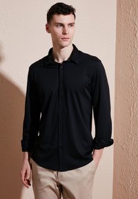 Zwarte blouse met lange mouwen, knoopsluiting aan de voorkant, klassieke kraag en opgerolde mouwen, gemaakt van zacht materiaal, gecombineerd met lichte broek.
