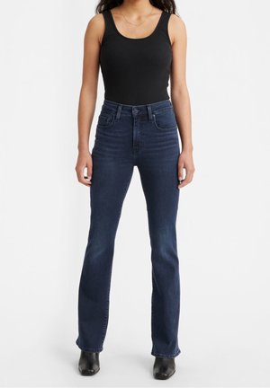Femme portant un jean bleu foncé taille haute, un haut noir sans manches et des bottines noires, debout devant un fond blanc uni.