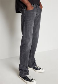 Jeans de mezclilla gris con un diseño de pierna recta, bolsillos frontales y un tenue desvanecido; combinados con zapatillas de lona en blanco y negro.