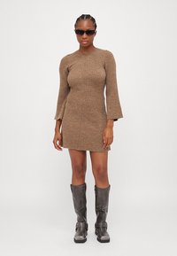 Robe en tricot côtelé marron avec de longues manches en cloche. Assortie à des bottes grises jusqu'aux genoux avec des boucles. Le mannequin porte des lunettes de soleil noires étroites.