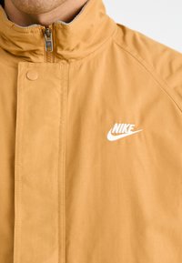 Giacca impermeabile gialla con colletto alto, dotata di una zip argentata e del logo Nike bianco ricamato sul petto. Texture liscia.