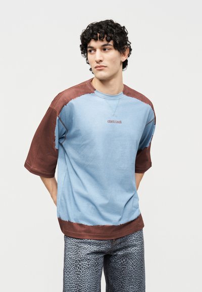 Νεαρός με σγουρά μαλλιά, που φοράει μπλε και καφέ κοντομάνικο πουκάμισο και ανοιχτό γκρι παντελόνι με σχέδια, κοιτάζει προς τα αριστερά του, σε απλό φόντο.