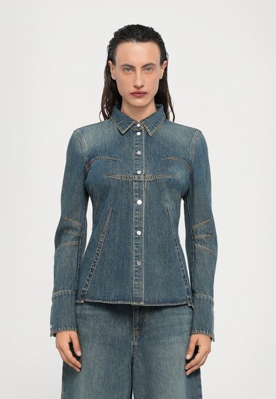 Chaqueta de mezclilla en azul oscuro con cierre de botones, cuello puntiagudo y detalles de costura. Presenta una forma ajustada y mangas largas.