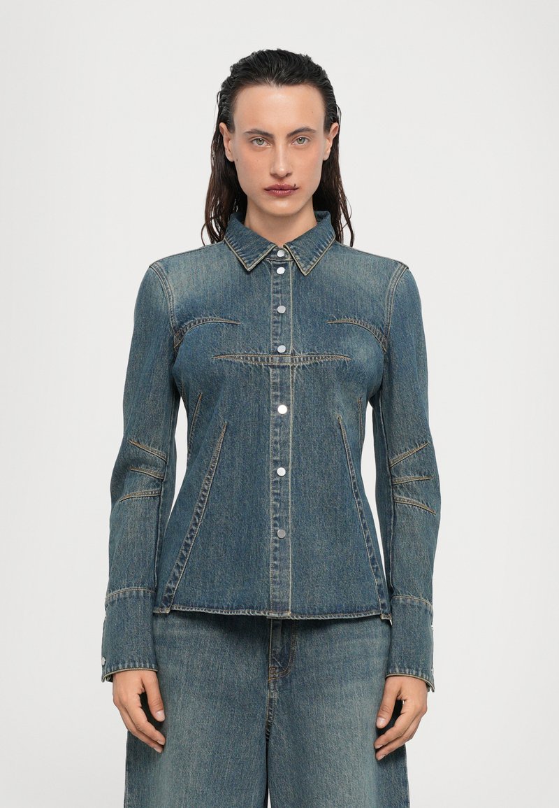 Giacca in denim di colore blu scuro con chiusura a bottoni, colletto a punta e dettagli di cucitura. Presenta una forma aderente e maniche lunghe.
