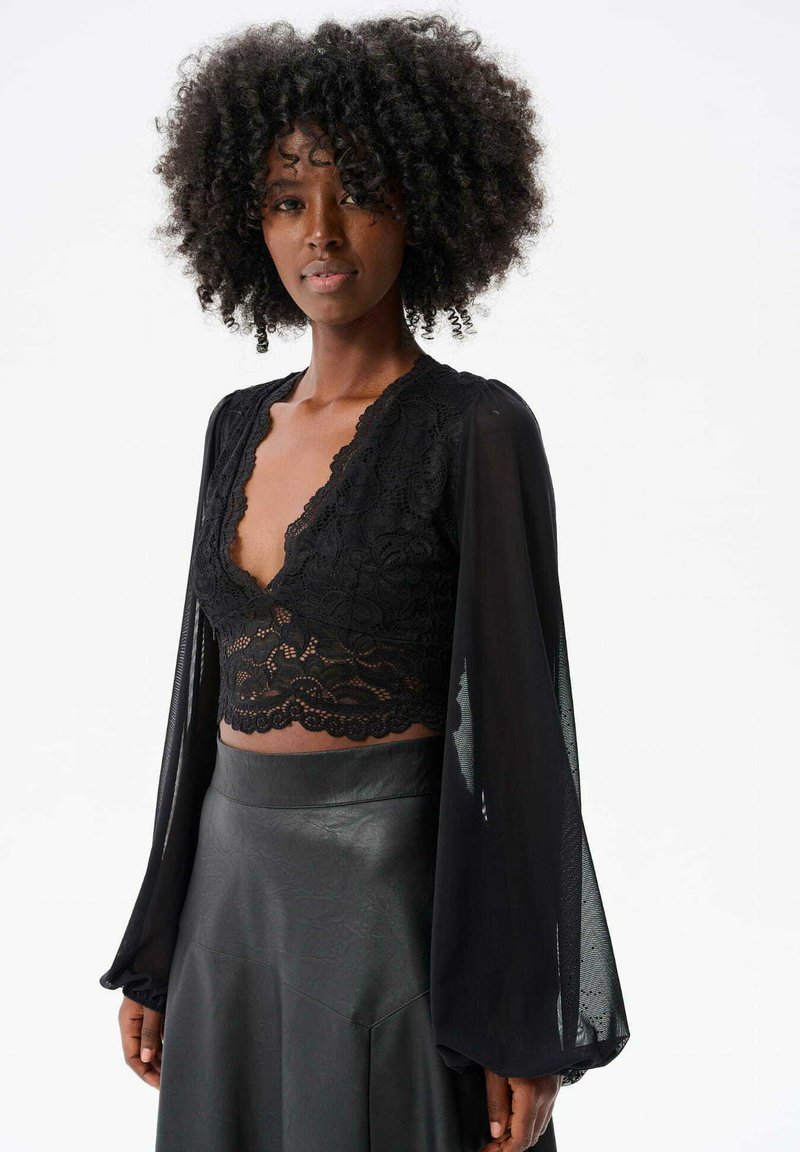 Czarna koronkowa bluzka z dekoltem w serek, długimi przezroczystymi rękawami i falbaniastym wykończeniem; zestawiona z dopasowaną czarną skórzaną spódnicą.