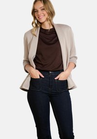Světle béžový blazer s otevřeným předním dílem a ohrnutými rukávy, vrstvený přes tmavě hnědý top, doplněný o vysoké džínové kalhoty tmavé barvy.