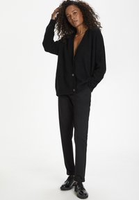 Black textured cardigan with a deep V-neck and two buttons, paired with black trousers and glossy black shoes. Neutral background.