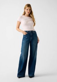 T-shirt rose pâle à manches courtes avec texte en relief, associé à un jean bleu foncé taille haute à jambes larges, présentant une texture denim lisse.