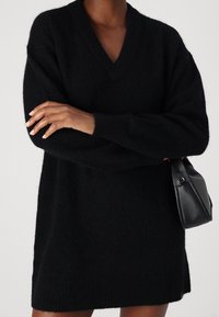 Robe pull noire oversize en maille avec col en V, poignets et ourlet côtelés. Associée à un petit sac à main en cuir noir.