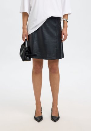 Woman wearing black leather skirt, white oversized top, black pointed heels, a silver bracelet, and holding a small black handbag.