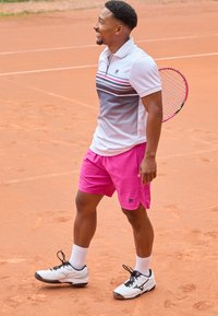 White polo shirt with gray and pink stripes, pink shorts, and white sneakers. Standing on a clay tennis court holding a racket.