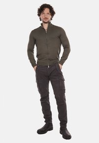 Pullover con zip di colore verde oliva, dal design aderente, abbinato a pantaloni cargo scuri con numerose tasche. Modello che indossa stivali neri.