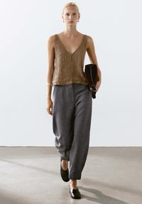 Brown textured sleeveless top with a v-neck and fringed hem, paired with loose gray trousers and black slip-on shoes. Black clutch in hand.