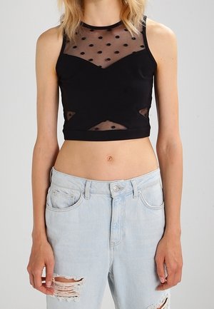 Kobieta w czarnym bezrękawniku typu crop top z przezroczystymi panelami w groszki i jasnoniebieskich podartych dżinsach, widoczna od ramion do bioder.