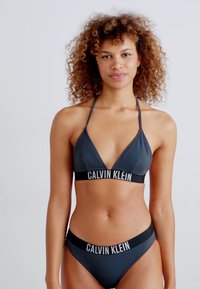 Set di bikini grigio scuro con top a triangolo e slip in stile hipster, entrambi con una banda nera che mostra la scritta 'CALVIN KLEIN' in testo bianco.
