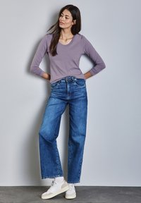 Maglietta lunga viola con scollatura a girocollo, abbinata a jeans blue a vita alta e scarpe da ginnastica bianche, in piedi su uno sfondo neutro.