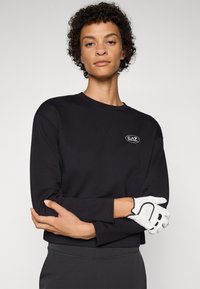 Μαύρος κοντός φούτερ από μαλακό ύφασμα, με λογότυπο στο στήθος. Το μοντέλο φοράει λευκά γάντια και στέκεται μπροστά σε έναν απλό φόντο.