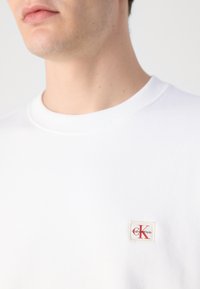 Suéter de algodón blanco con cuello redondo y detalles acanalados. Presenta un pequeño parche con el logo de Calvin Klein cosido en el lado izquierdo del pecho.