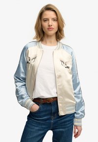 Giacca bomber in raso con sezioni crema e azzurro chiaro, gru ricamate, polsini a righe e chiusura con zip. Indossata sopra una t-shirt bianca.