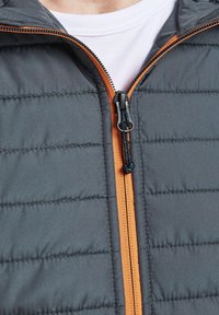 Grau gepolsterte Jacke mit orangefarbenem Reißverschluss, ausgestattet mit einer strukturierten Oberfläche und gestepptem Design, geeignet für Isolierung und den Einsatz im Freien.
