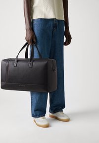 Black leather duffel bag with textured surface, dual handles, a subtle red stripe, and a zip closure. Worn with loose blue jeans and white sneakers.