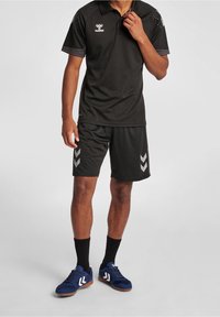 Svart atletisk polo med korta ärmar, textilstruktur och logotyp. Tillsammans med svarta shorts med silverfärgade chevrondetaljer. Blå atletiska skor.