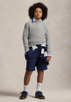 Bambino con capelli ricci che indossa un maglione grigio lavorato a treccia, pantaloncini blu, calzini bianchi e scarpe da ginnastica blu navy, in piedi con le mani in tasca.
