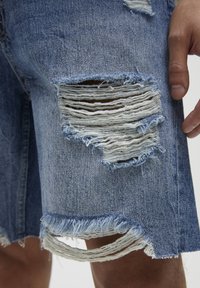 Shorts en denim bleu clair avec des détails usés et des bords effilochés. Présente des déchirures marquées et une texture rugueuse.