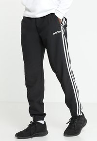 Osoba ve černých sportovních kalhotách Adidas s bílými pruhy a černých sportovních botách, stojící s rukama v kapsách.