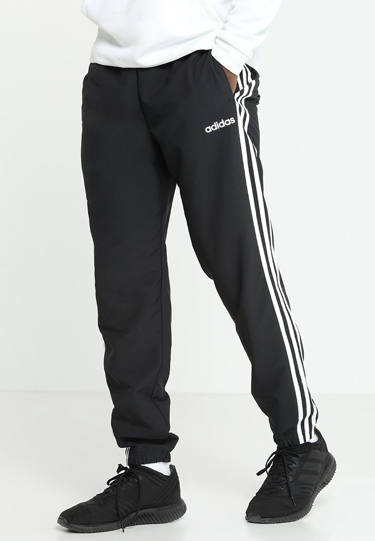 Osoba ve černých sportovních kalhotách Adidas s bílými pruhy a černých sportovních botách, stojící s rukama v kapsách.