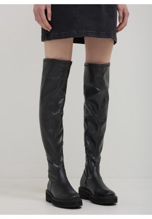 Persoon die zwarte lederen over-de-knie laarzen met grove zolen draagt en een korte zwarte rok, staand tegen een effen lichte achtergrond.