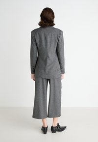 Sivina tailored obleka, ki vključuje strukturiran blazer z enim razporkom in širokimi hlačami. Teksturovan material s subtilnim vzorcem in gumbnimi akcenti.