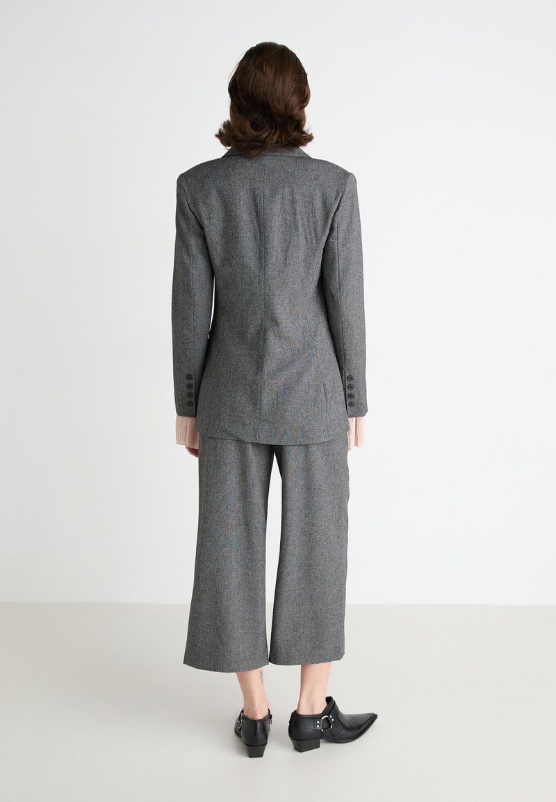 Traje gris a medida que presenta una chaqueta estructurada con una abertura trasera y pantalones de pierna ancha. Tejido texturizado con un patrón sutil y detalles de botones.