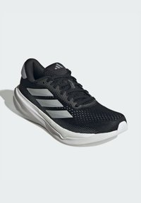 Chaussure de sport noire avec tige en maille, rayures argentées, col rembourré et semelle blanche texturée. Présente un design à lacets.