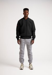 Giacca bomber nera trapuntata con polsini e vita a costine, abbinata a pantaloni della tuta grigi. Le calzature presentano un motivo beige e grigio.