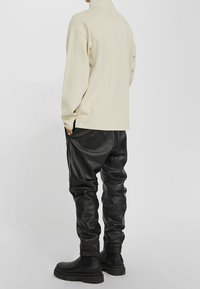 Béžový fleece svetr s vysokým límcem v kombinaci s černými koženými kalhotami a černými kotníkovými botami. Měkká textura, volný střih a boční kapsy.