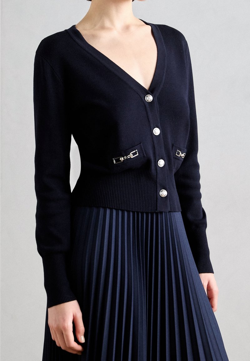 Cardigan bleu marine en tissu tricoté doux, doté d'un col en V, de six boutons et de deux poches avec des détails en métal argenté.