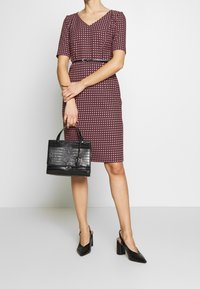 Robe jusqu'au genou, décolleté en V, de couleur bordeaux avec un motif géométrique blanc, taille cintrée, accompagnée d'un sac à main noir texturé et de talons slingback noirs.